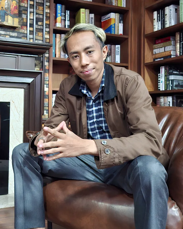 Aadarsh Shrestha in a brown jacket and navy check shirt, sitting forward in a leather armchair in front of bookshelves.