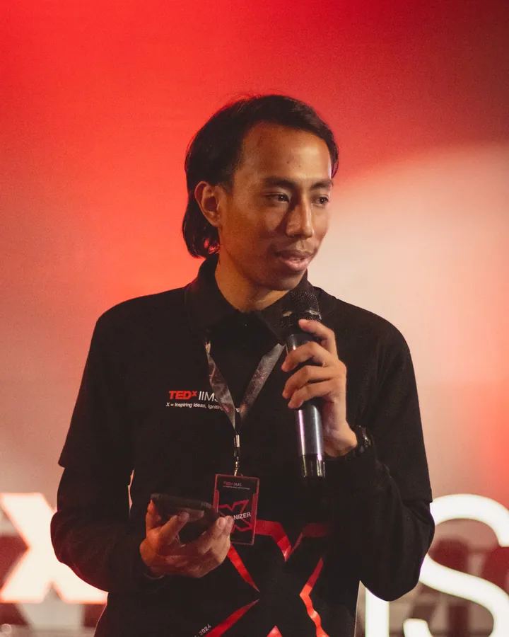Aadarsh speaking into a microphone on the TEDxIIMS stage, under red light.